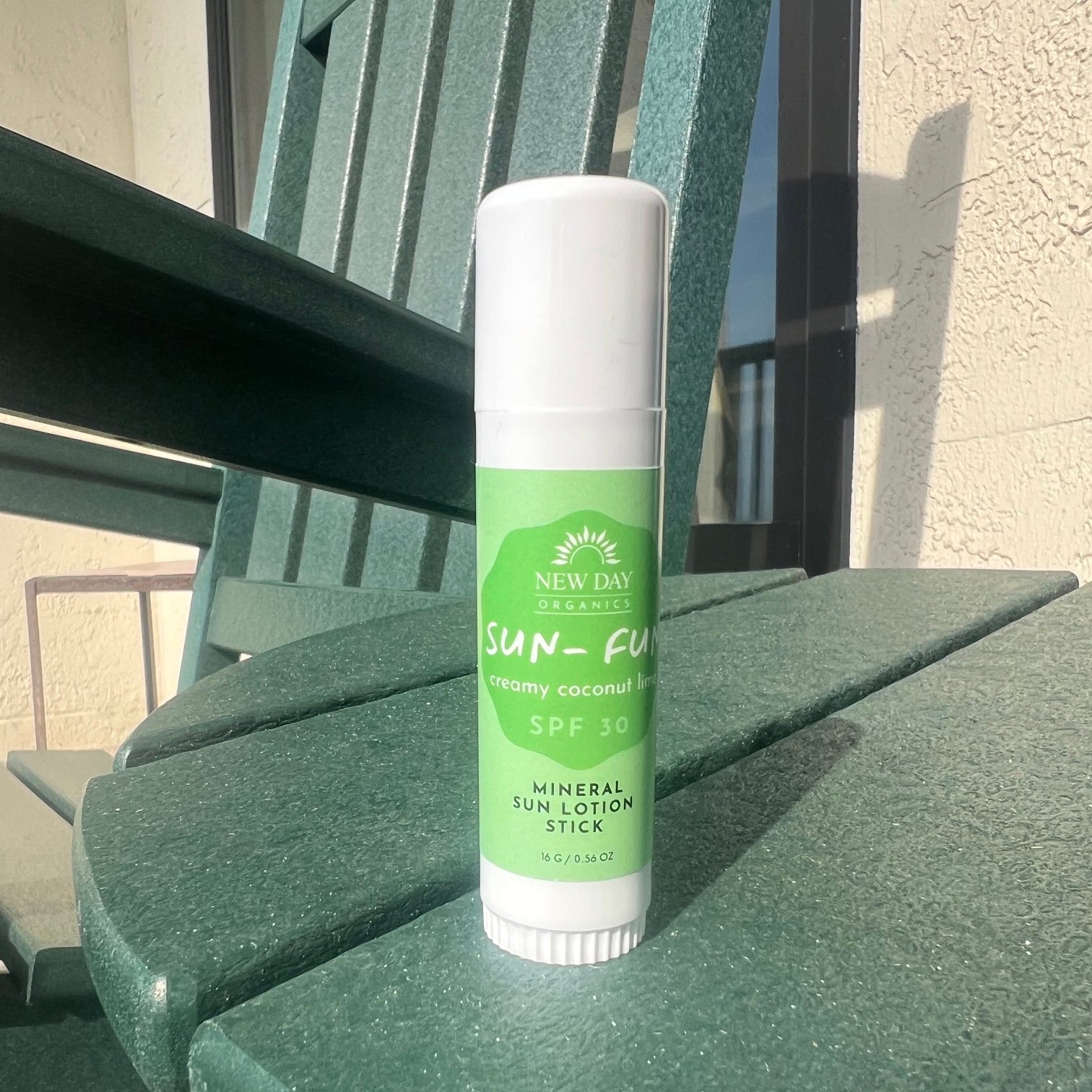 A white and green stick of New Day Essentials Sun-Fuel SPF 30 mineral sun lotion sits on a dark green outdoor patio chair in direct sunlight.