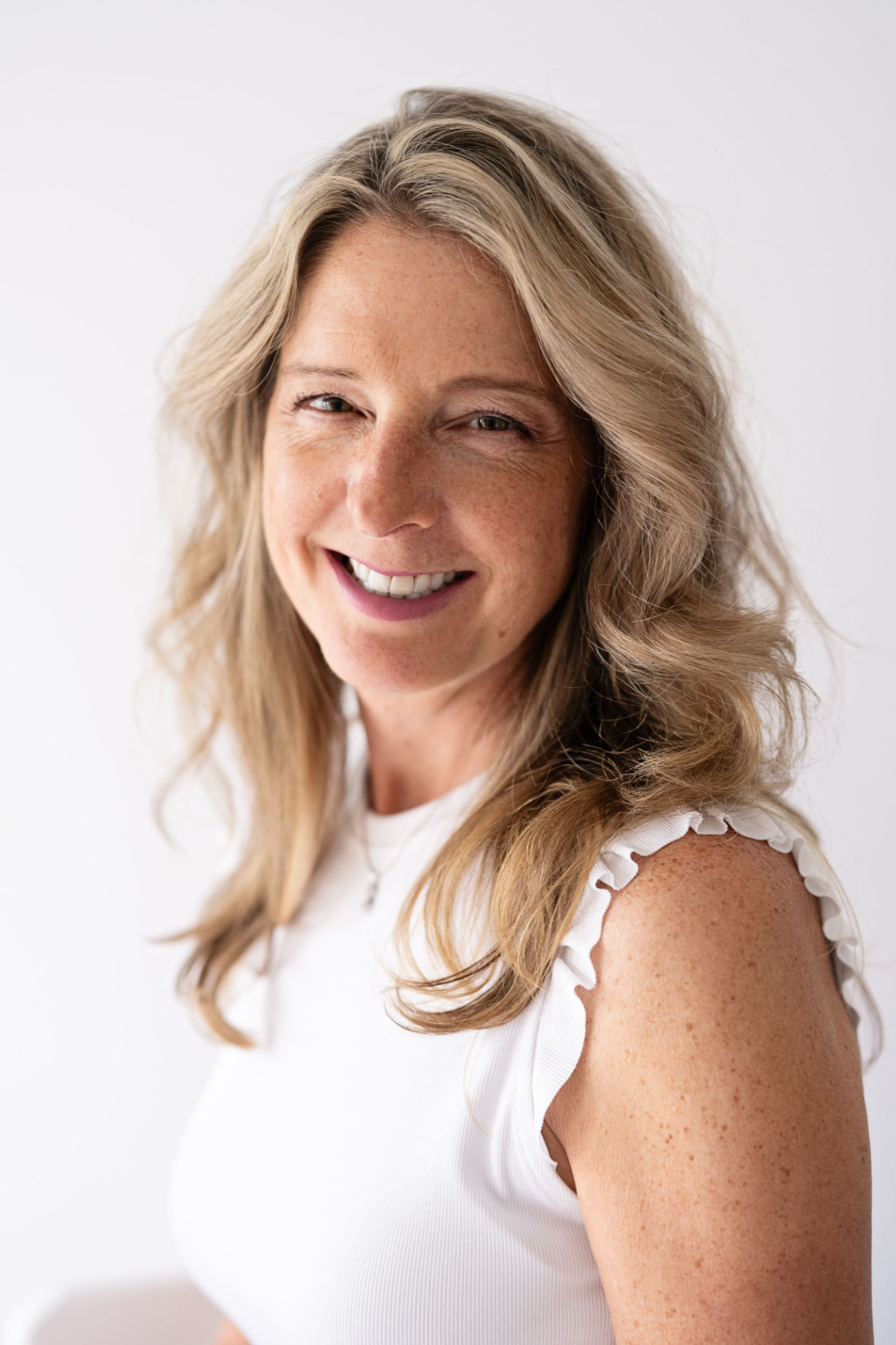 A smiling blonde woman with wavy hair, wearing a white sleeveless top with ruffled edges, photographed against a clean white background.