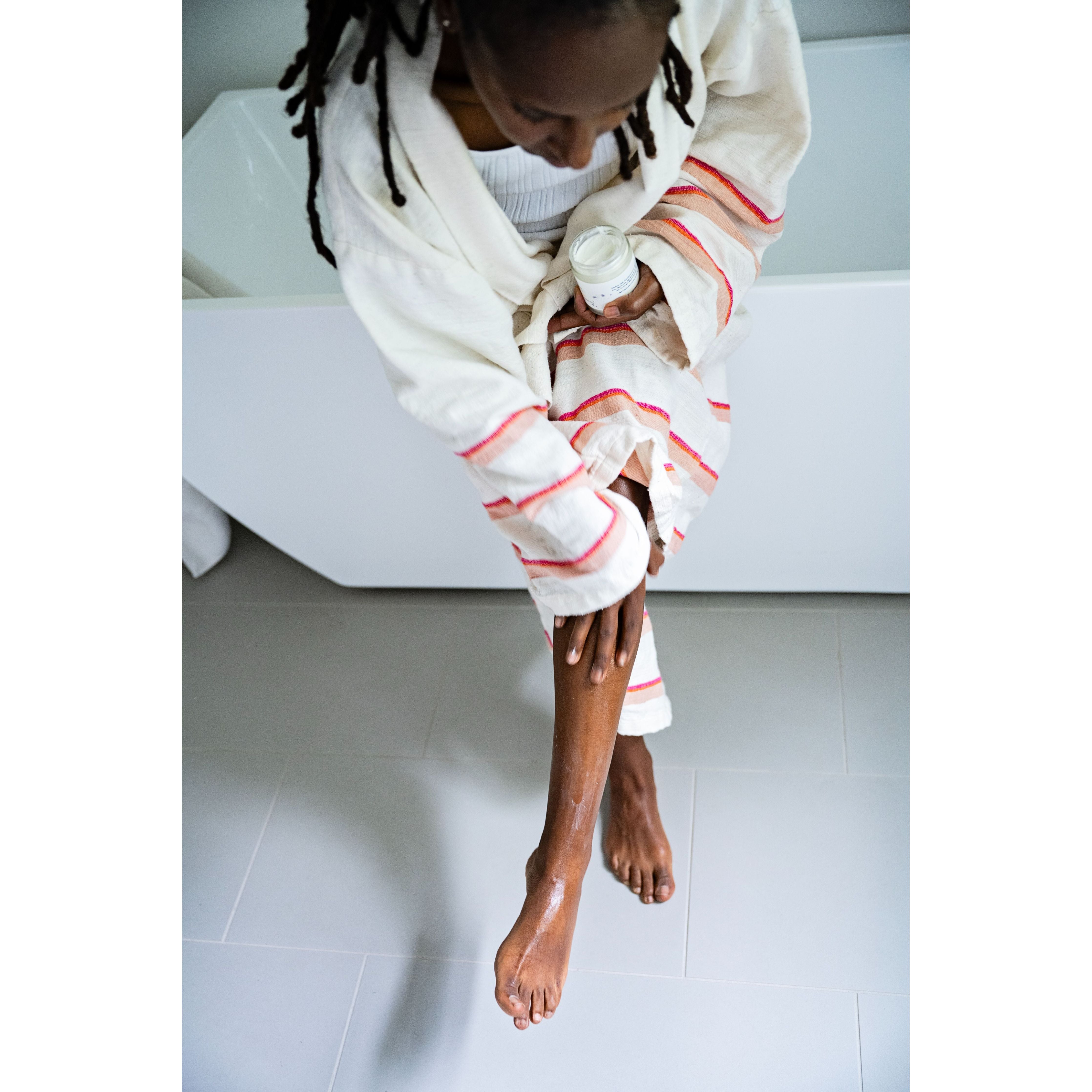 Woman sitting on a white bathtub with a towel wrapped around her, applying Whipped Shea Body Butter Souffle on her legs