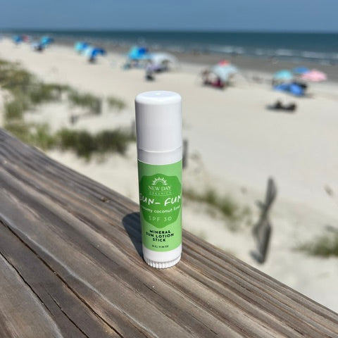 A small sunscreen stick labeled Sun-Fit SPF 50 sits on a wooden railing with a blurred beach scene in the background, including sand, ocean, and beach umbrellas.