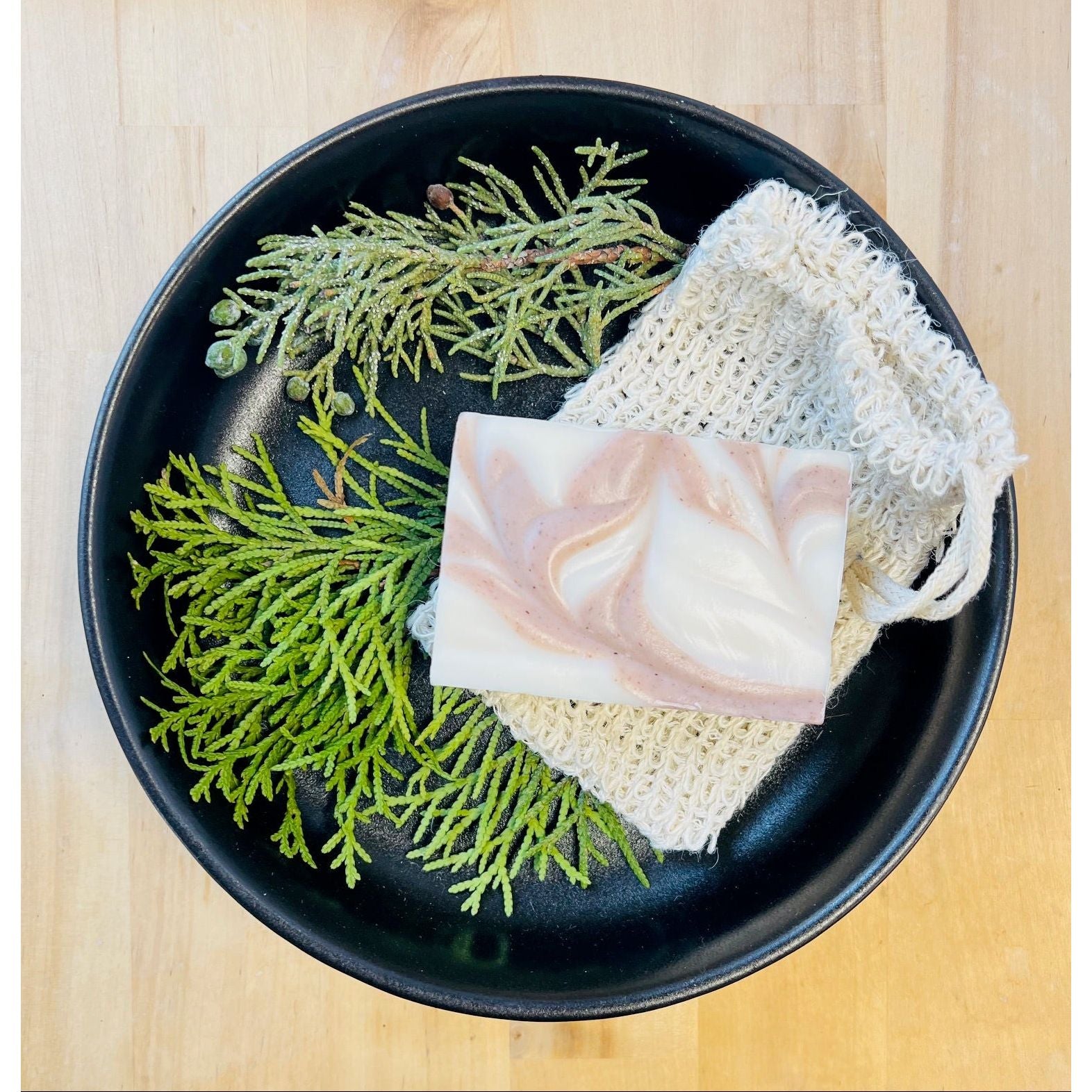 Black plate with pine and juniper, a bar of soap, and a loofah on a wooden surface.