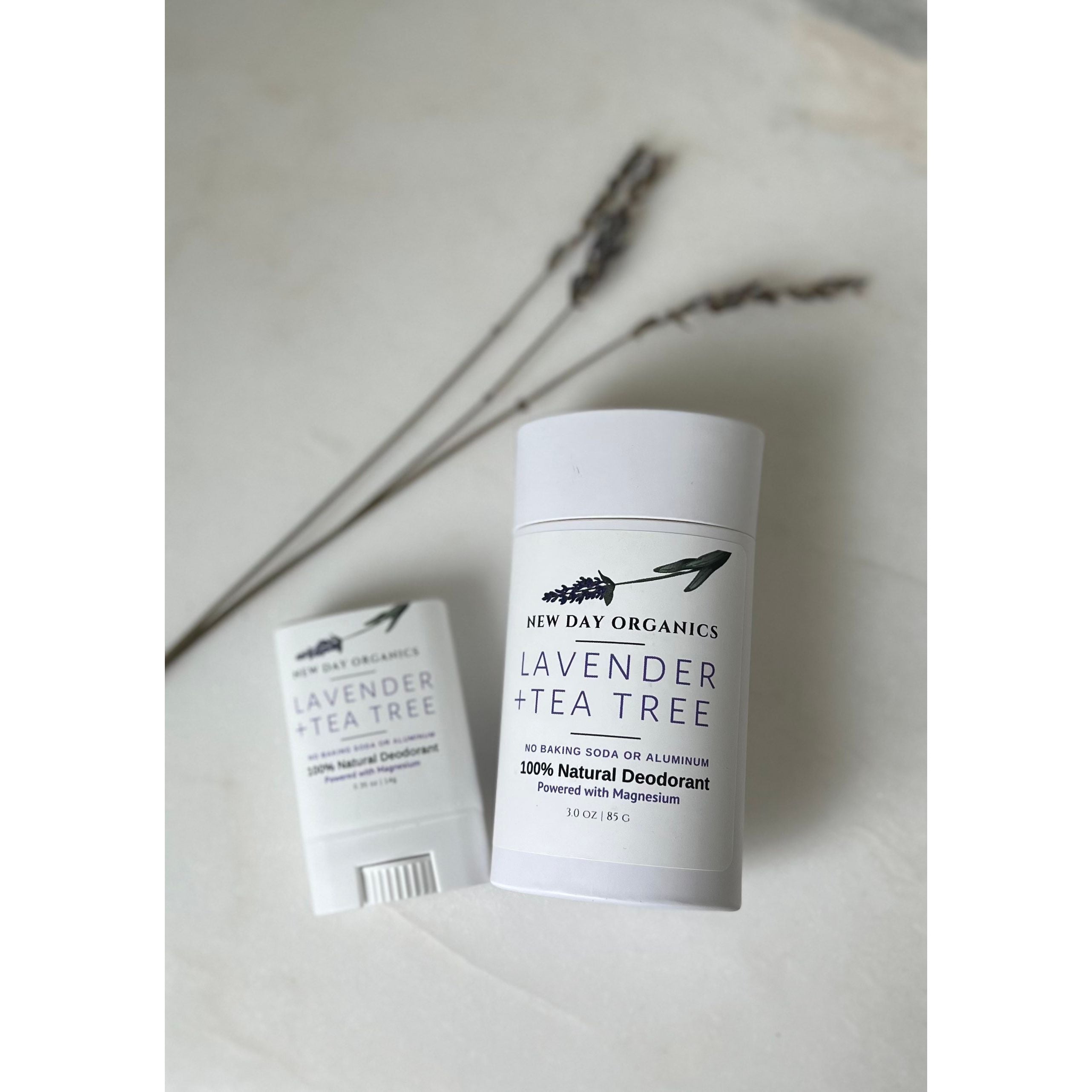 Two bottles of New Day Organics Lavender + Tea Tree natural deodorant on a light surface.
