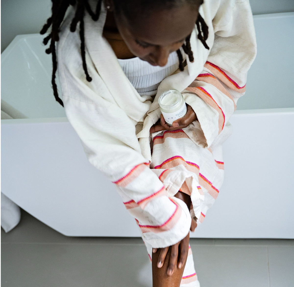 Woman sitting on a white bathtub with a robe, applying Whipped Shea Body Butter Souffle on her legs