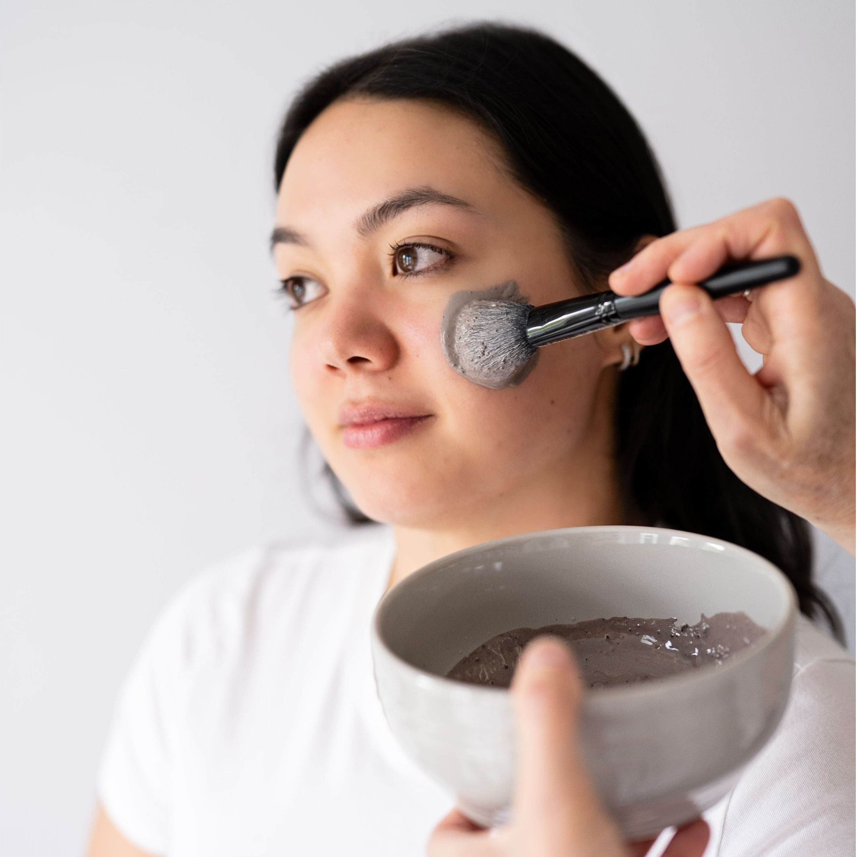 Woman applying a facial mask with a brush from a bowl against a plain background